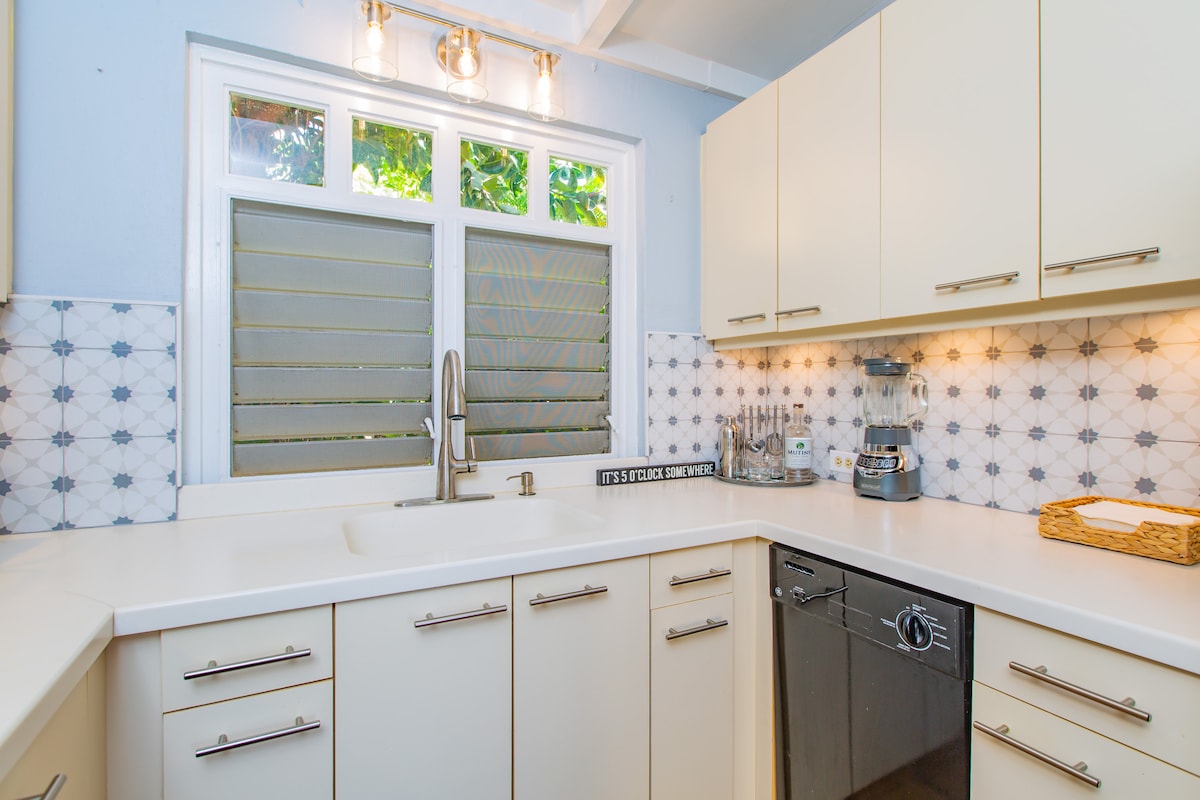 A modern galley kitchen is shown with light cabinetry and a spacious white countertop. Appliances include a stainless steel dishwasher and a coffee maker. Windows with louvered shutters allow natural light while showcasing views of the surrounding greenery.