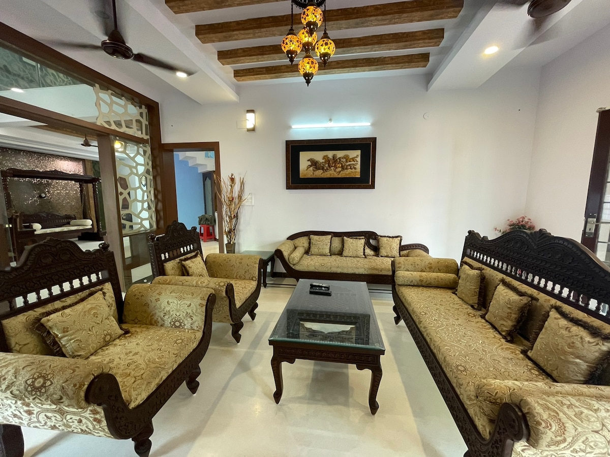 A spacious living room features elegant antique teak-wood sofas with intricate carvings, seating up to ten. A glass coffee table is centered in the room. Ambient lighting is provided by a ceiling fixture, and a framed artwork adds a cultural touch to the decor.