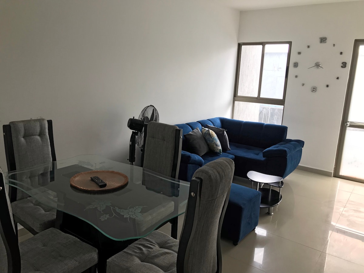 A modern living space is shown, featuring a dark blue sectional sofa adorned with decorative pillows. A glass dining table with six upholstered chairs is positioned nearby. Light streams through the large windows, illuminating the tiled floor, while a wall clock adds a subtle touch of elegance.