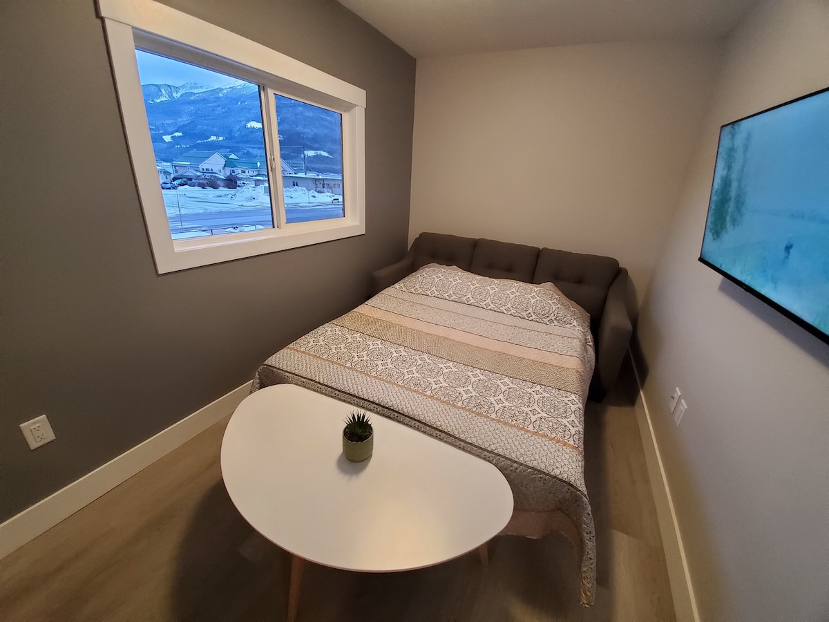 A well-appointed bedroom features a spacious bed topped with a patterned blanket. A sleek, low table is placed in front of the bed under a window, which offers views of the outdoor scenery. A wall-mounted television is positioned opposite the bed.