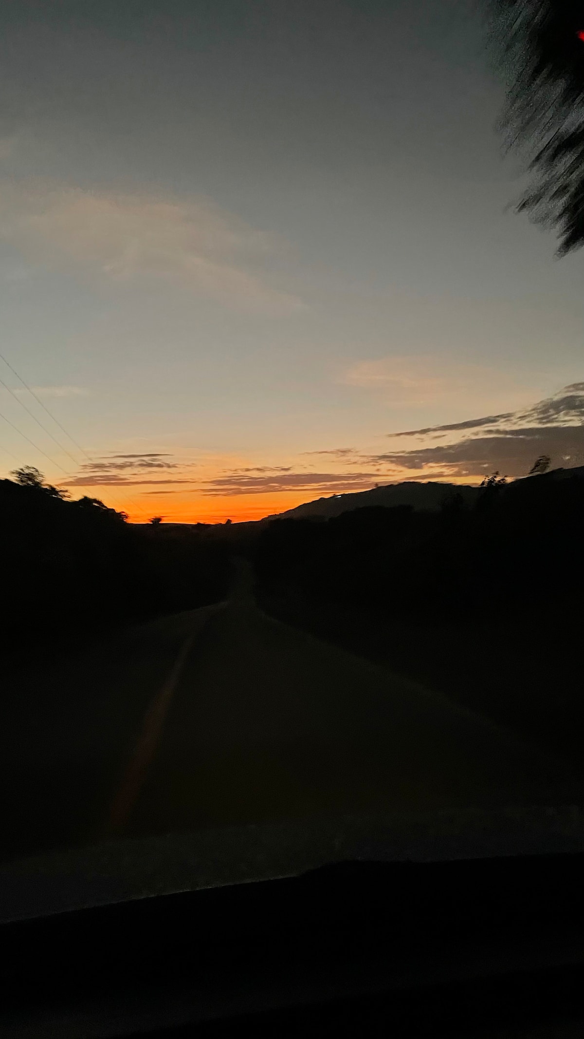 A scenic road stretches into the distance, leading toward a horizon illuminated by a warm sunset. The sky transitions from deep blue to vibrant orange hues, while silhouettes of trees and mountains create a tranquil backdrop.