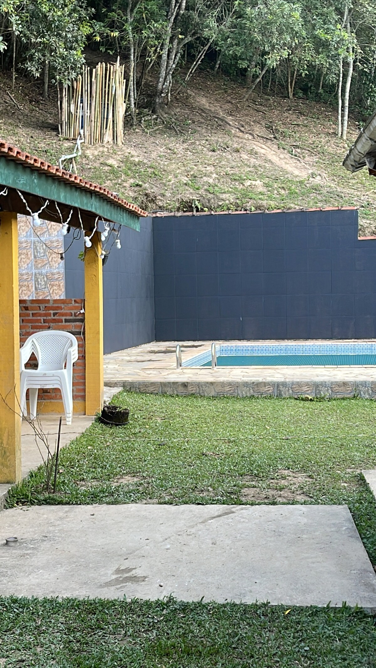 A serene outdoor area features a swimming pool surrounded by a stone deck, with a grassy lawn bordered by trees. A small patio area includes a white plastic chair positioned on a concrete surface, showcasing a blend of natural and built environments.