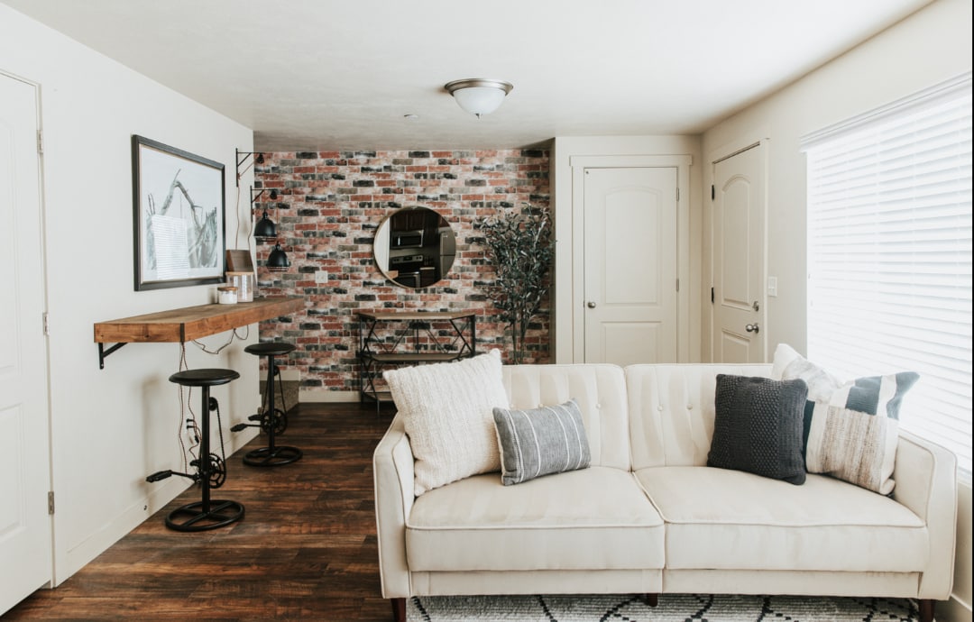 A modern living room features a light-colored sofa with decorative cushions. Exposed brick accents are present on one wall, while a round mirror reflects the space. A small dining area with stools and a wooden bar sits adjacent to the couch, complemented by natural light from a nearby window.