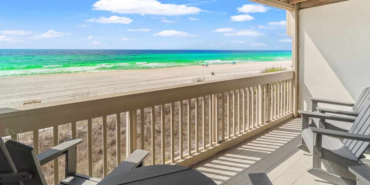 A covered deck is visible, featuring two rocking chairs and a table with chairs. The oceanfront view reveals vibrant turquoise waters and sandy beach, with a few distant beachgoers. The space is designed for relaxation, providing a serene backdrop for outdoor dining or drinks.