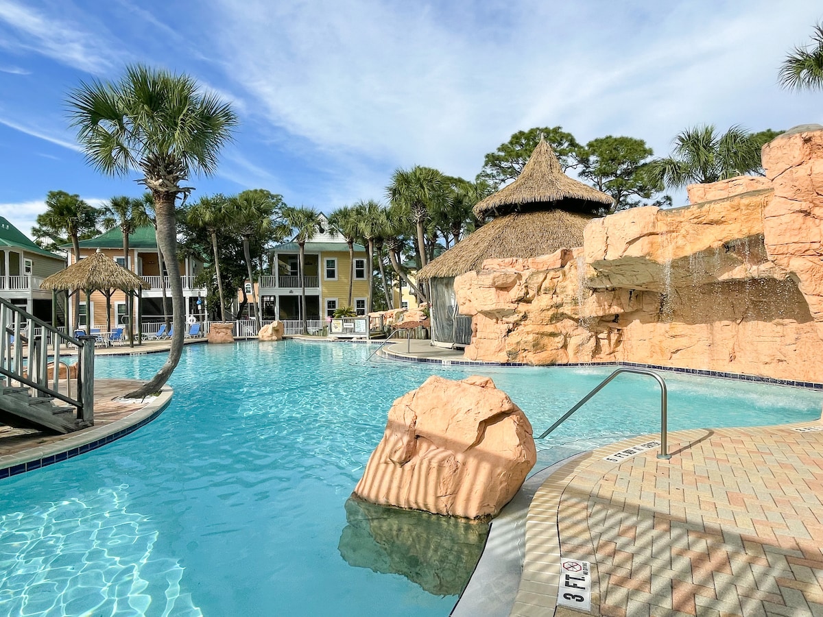 A landscaped lagoon pool is surrounded by palm trees and rock formations, featuring a waterfall cascading down from a natural stone structure. A thatched-roof gazebo is visible in the background, while colorful townhouses are arranged in a picturesque line overlooking the water.