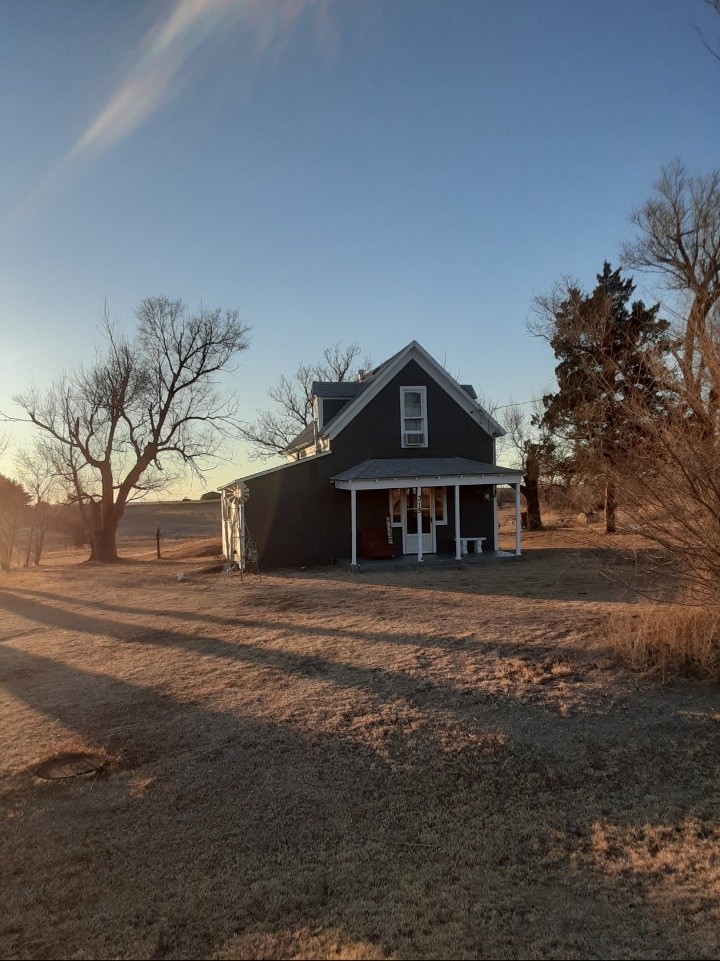 A two-story house is positioned on a spacious plot, surrounded by open land. The exterior features dark siding and a gabled roof, while the porch is accessed by a few steps. Trees and brush are visible in the background under a clear sky.