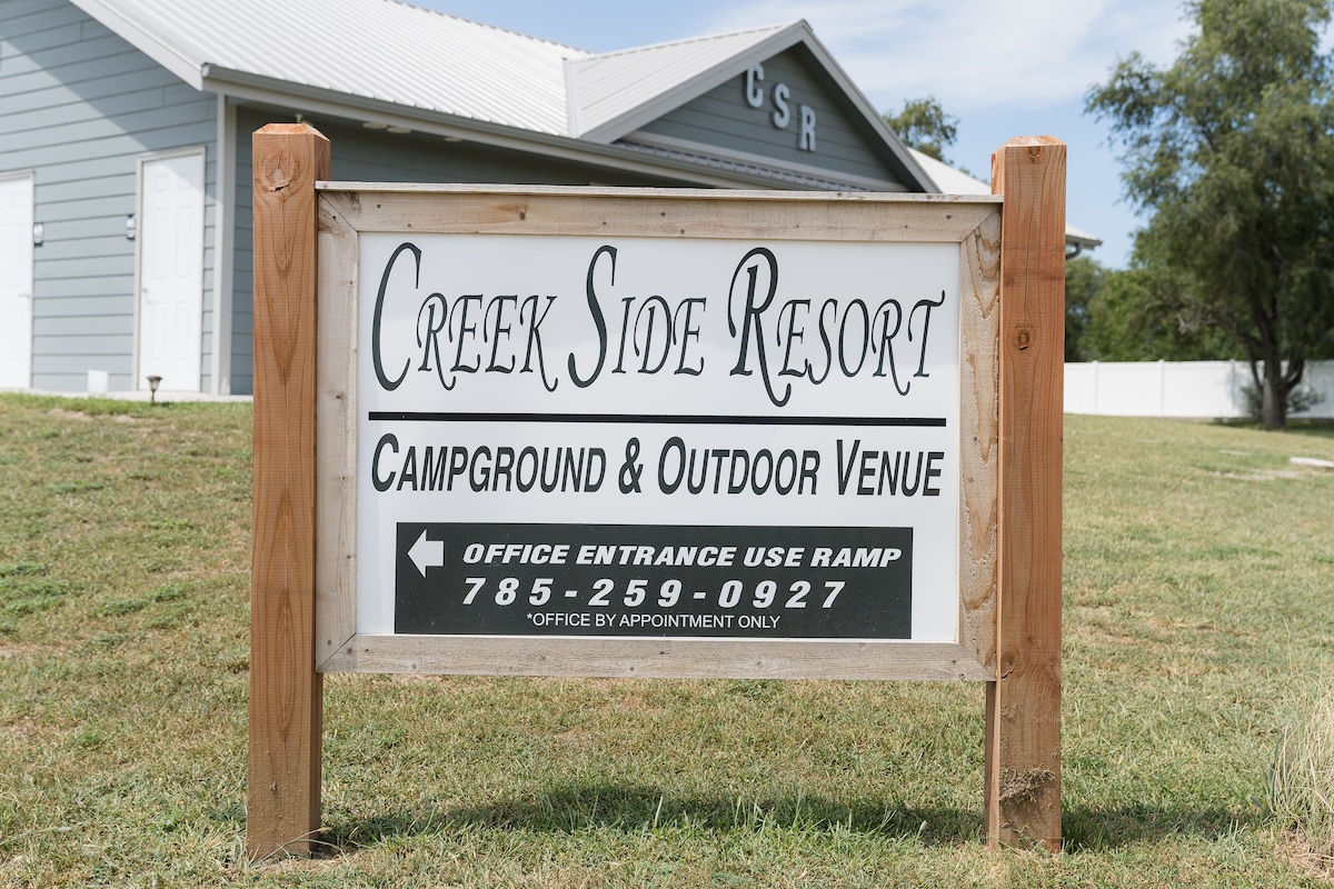 A wooden sign displays the name 'Creek Side Resort' along with details about camping and outdoor venue options. The sign is positioned on grassy ground, with a building visible in the background. The office entrance is indicated to use a ramp.