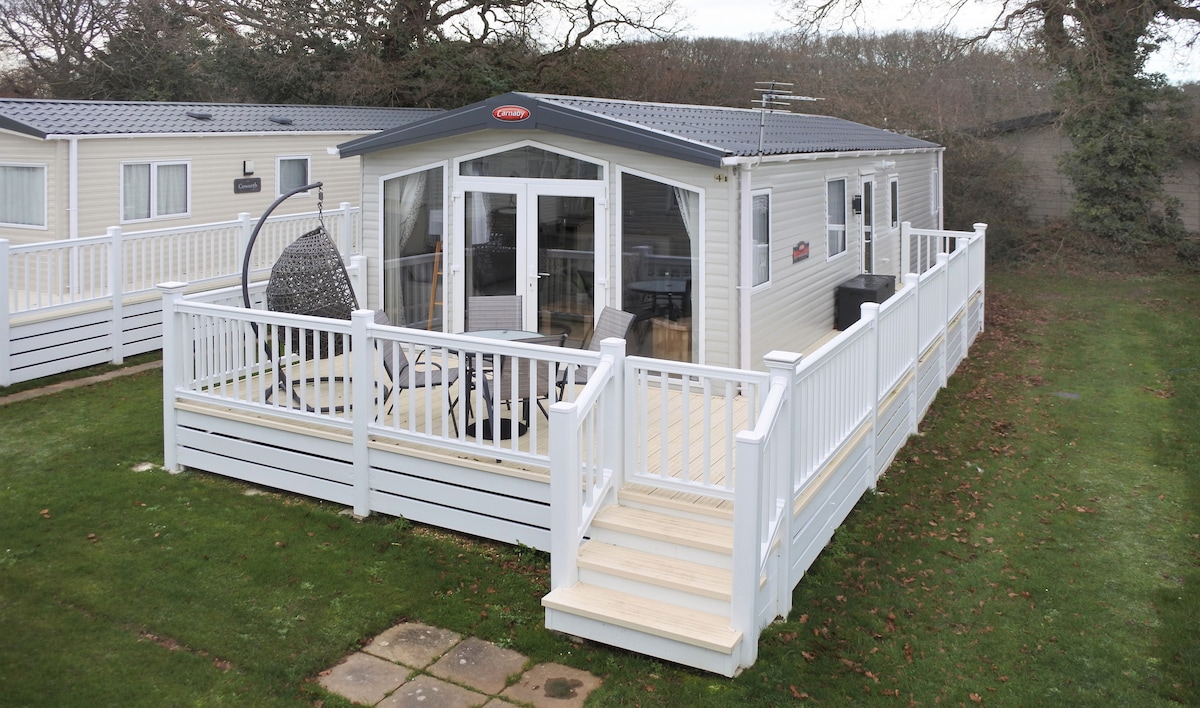 A modern caravan is situated on a spacious wooden deck, surrounded by greenery. The exterior features large glass doors that invite natural light into the interior. A contemporary hanging chair adds a relaxed touch, while an open area promotes outdoor leisure.