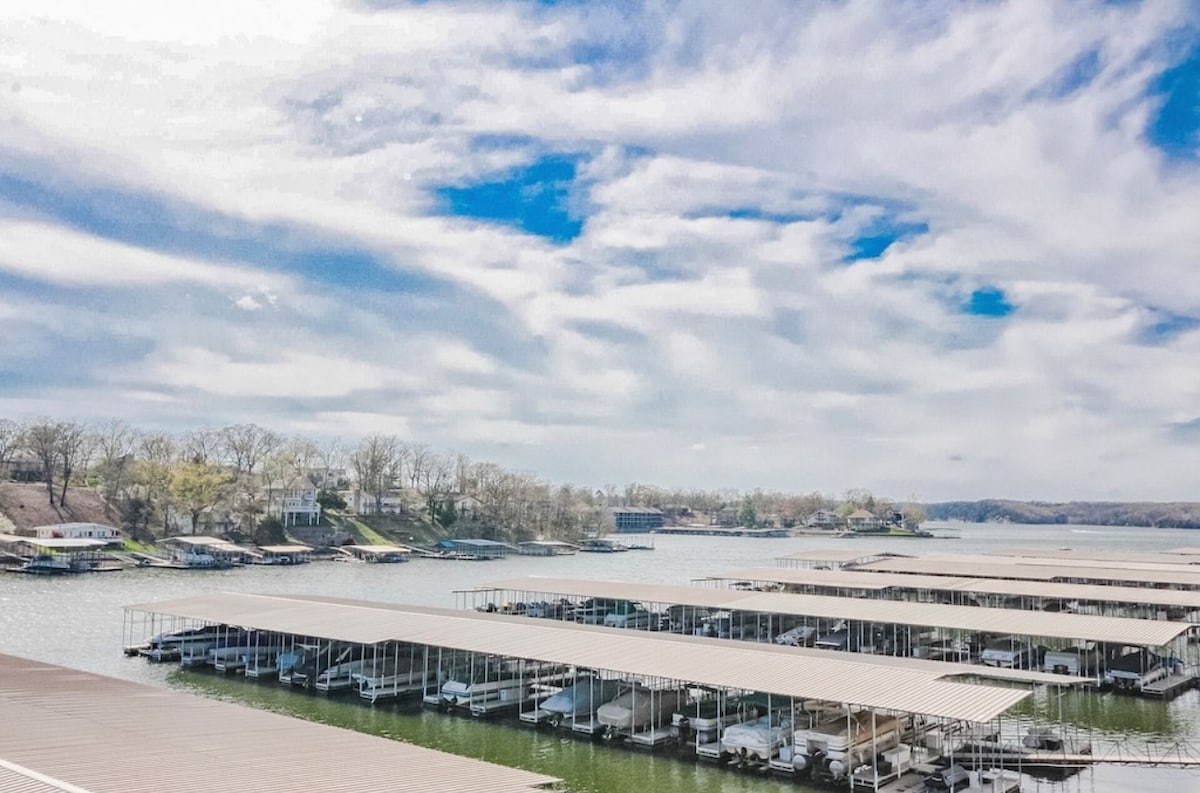 A scenic view of a marina featuring multiple boat docks. The water is calm, reflecting the sky filled with soft, white clouds. Tree-lined shores are visible in the distance, enhancing the serene waterfront setting.