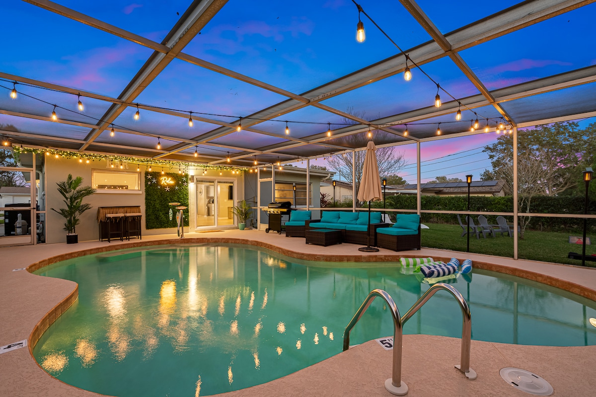 A serene outdoor space features a heated saltwater pool with gentle lighting, surrounded by a screened enclosure. Comfortable seating areas are arranged nearby, with a covered space for relaxation and an outdoor bar in the background, creating an inviting atmosphere for leisure and gatherings.