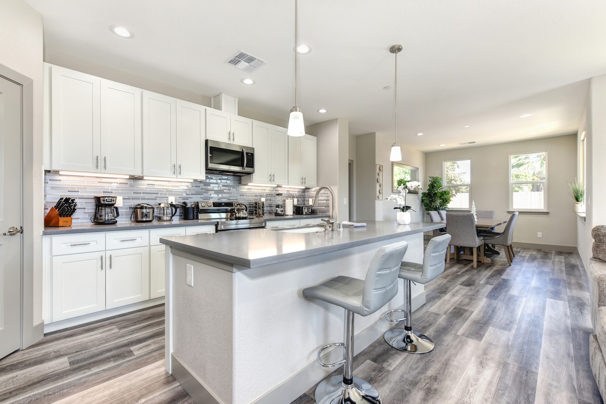 A modern kitchen is equipped with stainless steel appliances and white cabinetry, complemented by a large island with bar seating. The open layout connects to a dining area featuring a large table and chairs. Natural light enters through multiple windows.