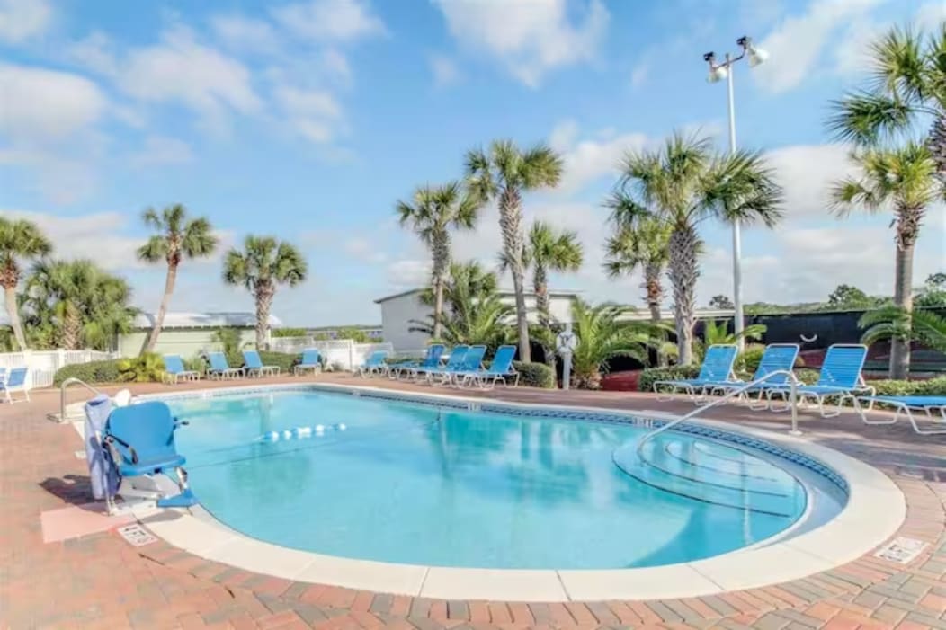 A serene pool area is surrounded by palm trees, with lounge chairs arranged neatly around the perimeter. The pool features clear, inviting water and a gentle slope leading into the shallow end. The sky is bright and clear, providing a pleasant atmosphere for relaxation.