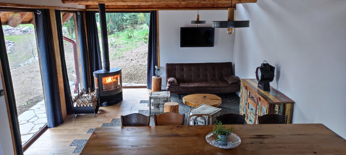 An open living area features large windows allowing natural light to fill the space. A comfortable seating arrangement includes a brown sofa and natural wood accent pieces. A stylish stove provides warmth, while a wooden table sits center, surrounded by chairs and decorative elements.