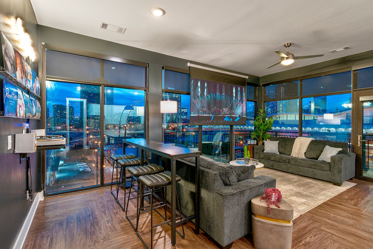 A modern living space is showcased with large panoramic windows revealing a city skyline. The area features a comfortable gray sofa, a long dining table with bar stools, and a cozy rug. Ambient lighting enhances the inviting atmosphere of the room.