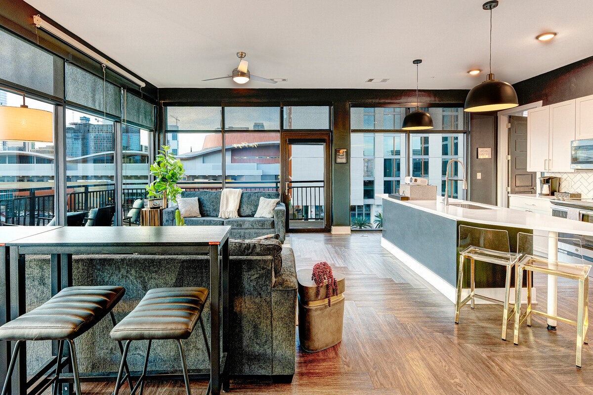 A spacious living area features a comfortable sectional sofa positioned near large floor-to-ceiling windows, offering ample natural light. A modern kitchen with sleek finishes is seen in the background, complemented by minimalist bar stools around a stylish counter.