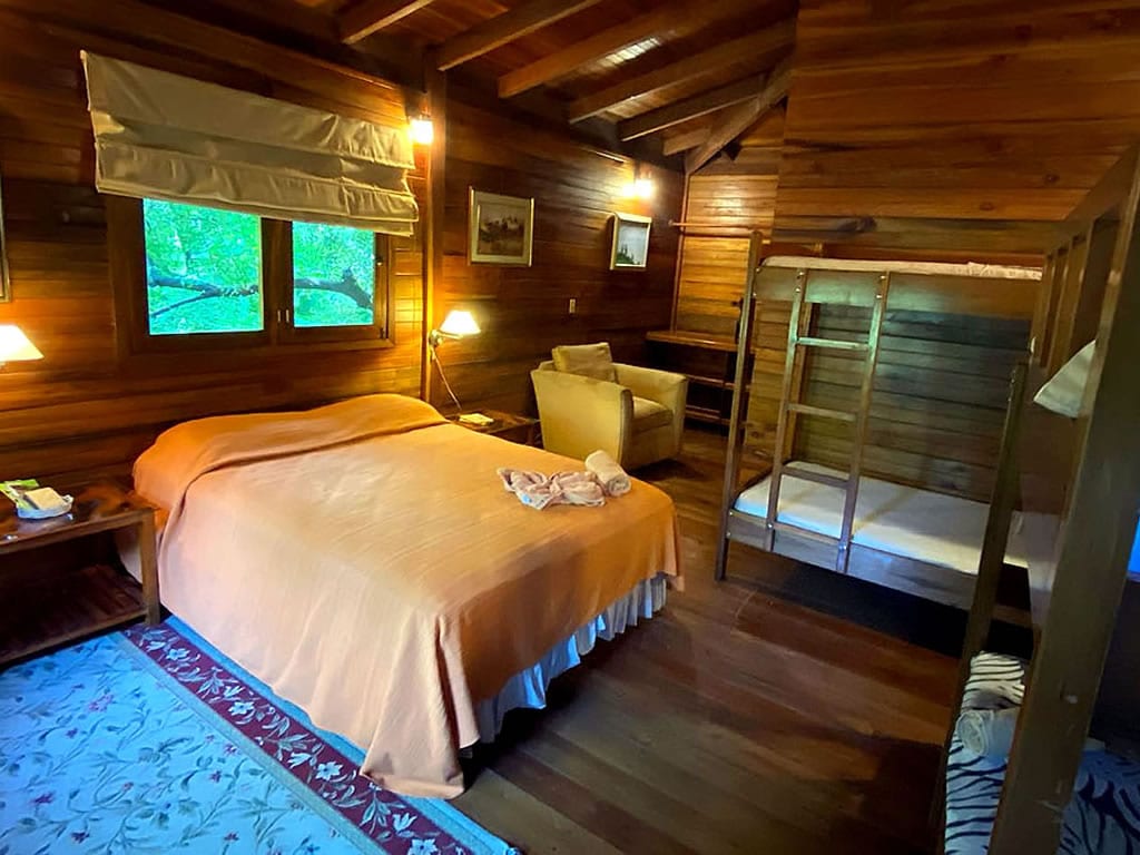 A rustic room is featured, showcasing wooden walls and beams. The layout includes a spacious bed covered with an orange blanket, a cozy seating area with an armchair, and a bunk bed. Natural light filters through windows adorned with light curtains.