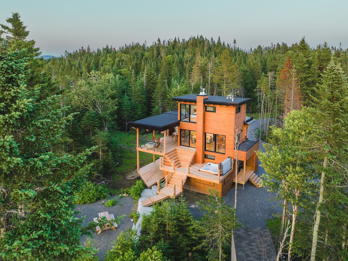 The micro-house is nestled among greenery, showcasing a modern wooden exterior. Multiple terraces extend from the structure, with a hot tub visible on the deck. The surrounding lush forest provides a serene natural backdrop, enhancing the home's integration into the landscape.