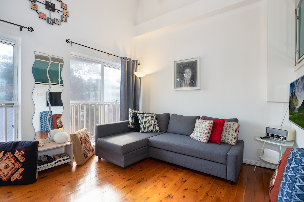 A modern living area features a grey sectional sofa adorned with colorful cushions. A mirrored wall and a striking wall art piece add visual interest. Natural light is invited in through large windows, complementing the warm wooden flooring.