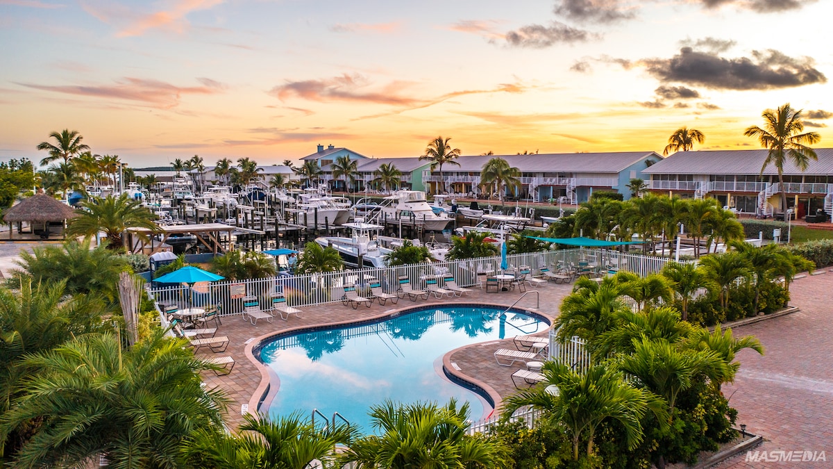 A view of the marina and pool area showcases multiple boats moored along the dock. An inviting pool is surrounded by lounge chairs, with tropical landscaping adding greenery. The sky exhibits soft hues of orange and pink, reflecting the peaceful atmosphere of the complex.