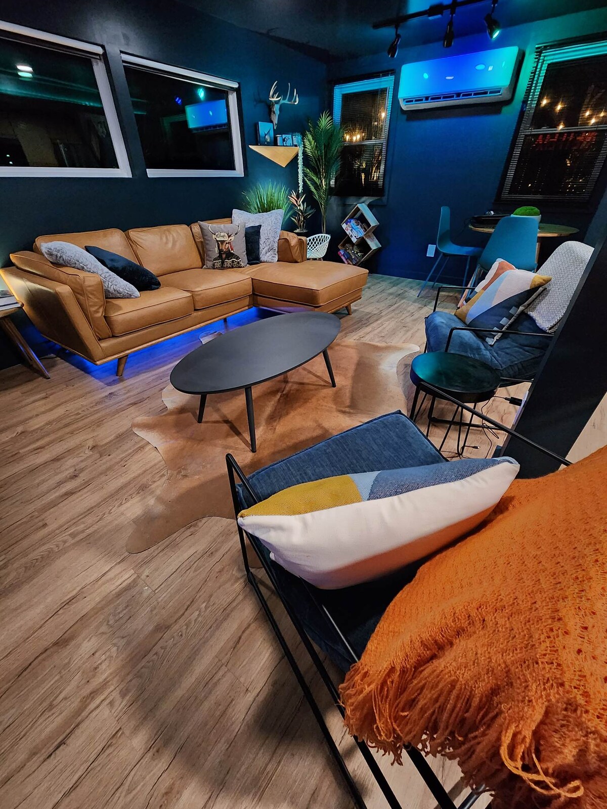 A modern living area features a stylish leather sectional sofa paired with a round coffee table. Colorful accent chairs are arranged around a soft area rug. Large windows frame the space, allowing natural light to enhance the inviting atmosphere.