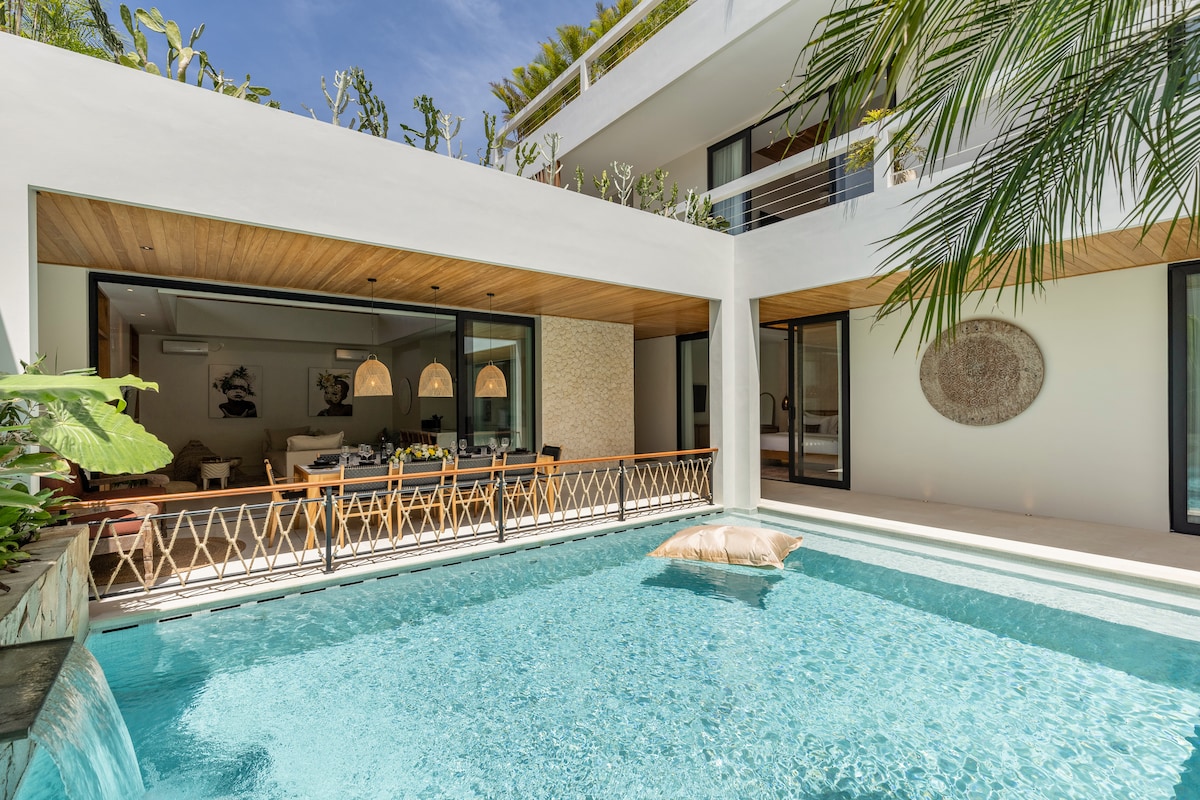 A serene outdoor area features a clear swimming pool, inviting relaxation. The pool is bordered by modern architecture and lush greenery, with sunlight reflecting off the water's surface. Sliding glass doors provide a view into a spacious dining and living area, enhancing the open atmosphere.