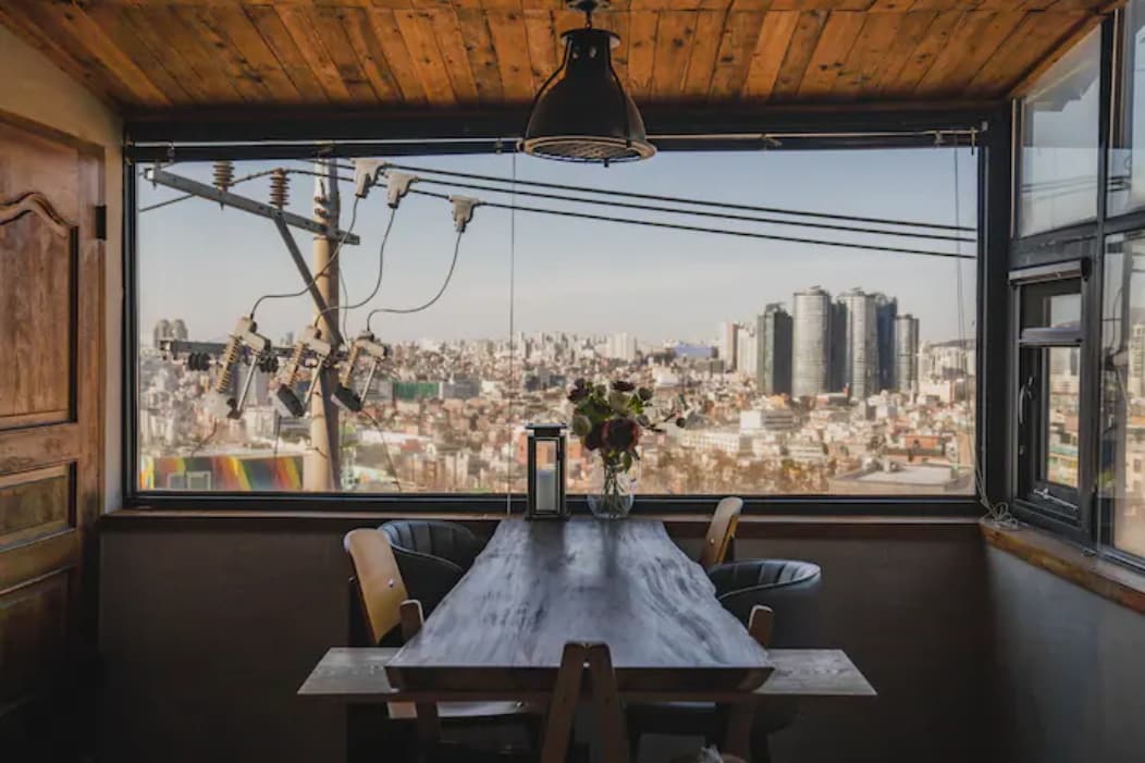 A dining area features a wooden table surrounded by chairs, set against a large window that offers views of the city skyline. Natural light fills the space, highlighting a floral centerpiece and a coffee maker on the table.
