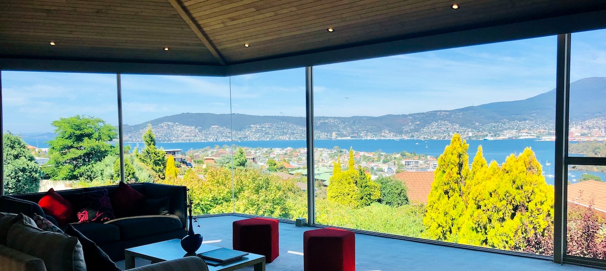 A spacious lounge area features large floor-to-ceiling windows that frame panoramic views of the Derwent River and surrounding hills. Comfortable seating is arranged thoughtfully, with vibrant accent stools positioned nearby. Natural light fills the space, highlighting the natural surroundings outside.