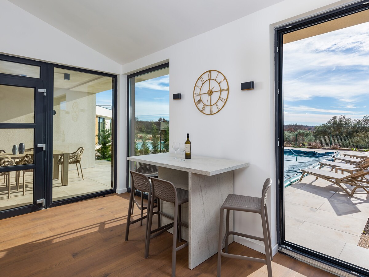 A dining area is displayed with a modern table and four high-backed chairs. Large glass doors open to the outdoor space, revealing a sunlit terrace and swimming pool. Minimalist decor is accentuated by a wall clock and a bottle of wine, inviting a relaxed atmosphere.