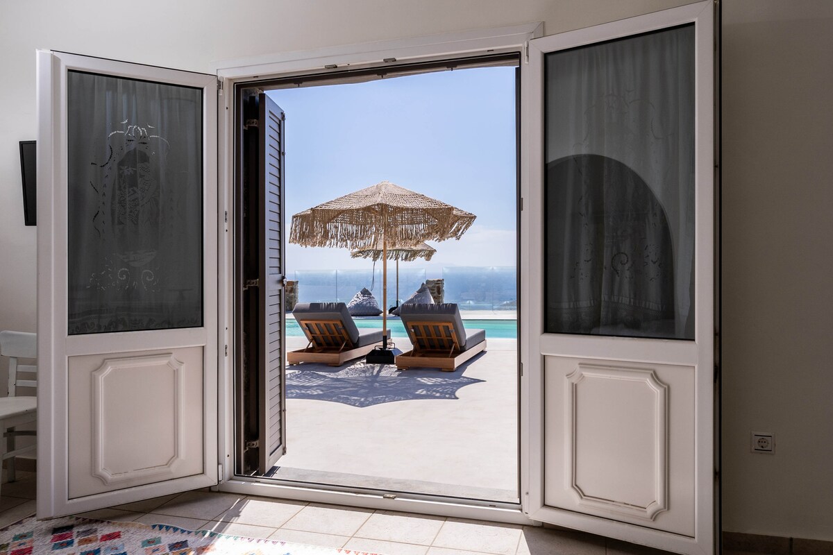 Double doors open to reveal a terrace with two lounge chairs and a sun umbrella, overlooking a shimmering sea. The sunny outdoor space features stunning views and invites relaxation.