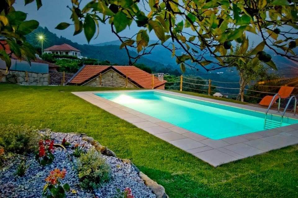 A private pool is set against a backdrop of rolling hills and carefully landscaped greenery. Surrounding the pool, vibrant flowers and smooth stones create a serene outdoor space, while illuminated water reflects the calming ambiance of the evening sky.