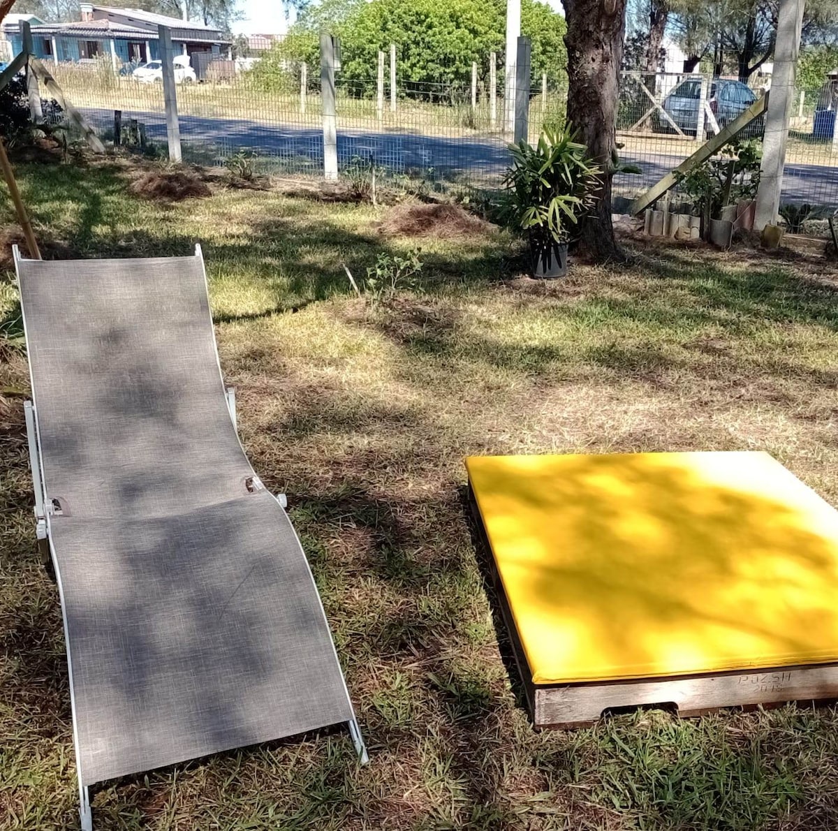 A sunlit outdoor area features a gray lounge chair next to a bright yellow platform. The grass is well-maintained, with surrounding greenery and trees providing shade. A tranquil atmosphere is created, ideal for relaxation or enjoying the pleasant outdoor environment.