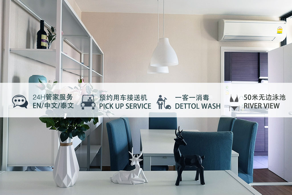 A modern dining area features a white table surrounded by six chairs. A decorative vase and a figurine of a deer are placed on the table. A pendant light hangs above, and a glimpse of a river view is visible through the nearby window.