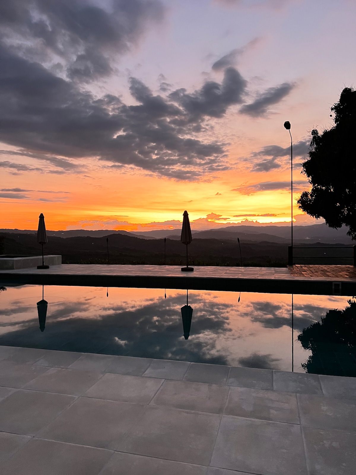 A tranquil pool reflects a vibrant sunset, with colorful clouds illuminated by soft hues. Surrounding features include umbrellas positioned on the pool deck against a backdrop of distant hills, creating a serene environment for relaxation.