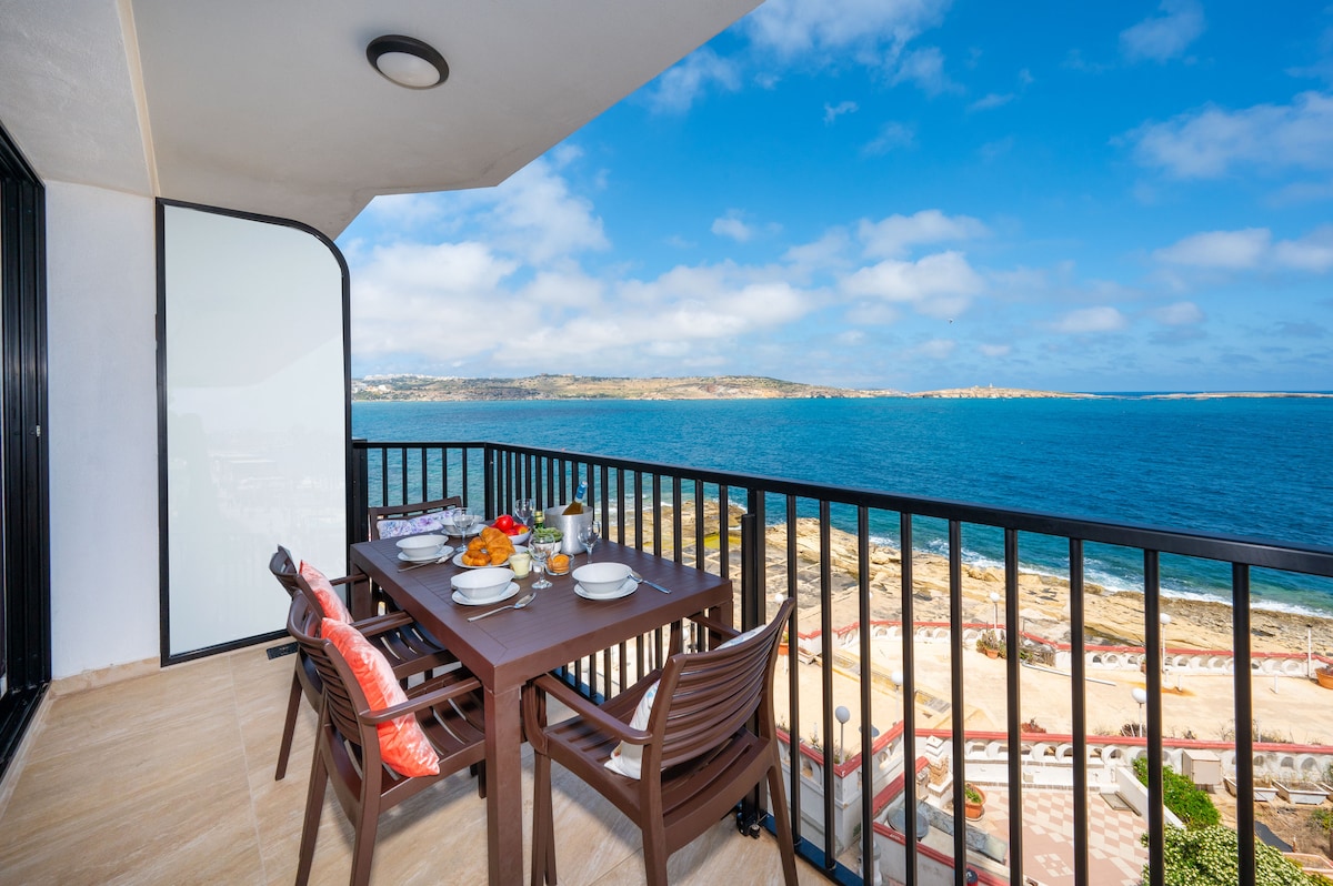A corner terrace is featured, overlooking the sea and islands. A dining table is set with food and drink, surrounded by comfortable chairs. The space is complemented by a railing providing unobstructed views of the water and coastline under a bright, partly cloudy sky.