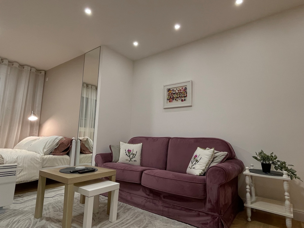 A cozy seating area features a plush purple sofa adorned with floral cushions. A light-colored coffee table sits in front, accompanied by a small white side table. The space is well-lit with recessed ceiling lights, and a comfortable bed is visible in the background.
