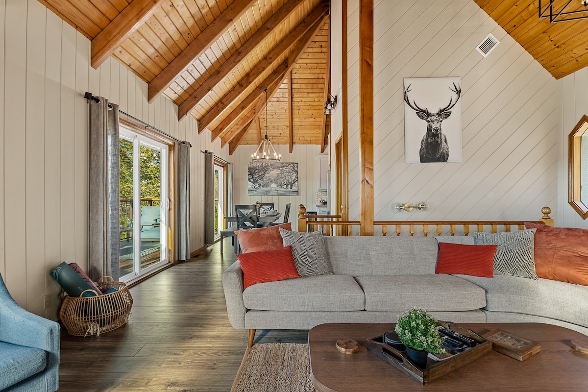 A spacious living area showcases high, vaulted wooden ceilings with exposed beams. A comfortable gray sofa is adorned with orange cushions, positioned near a coffee table. Large windows allow natural light to fill the room, offering views of the outdoor space and surrounding nature.
