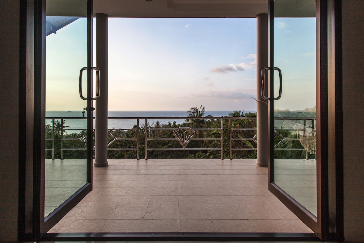 A spacious balcony opens up to a stunning view of the sea and surrounding greenery. Double doors frame the entrance, inviting natural light into the area. The flooring is tiled, contributing to a clean and modern aesthetic.