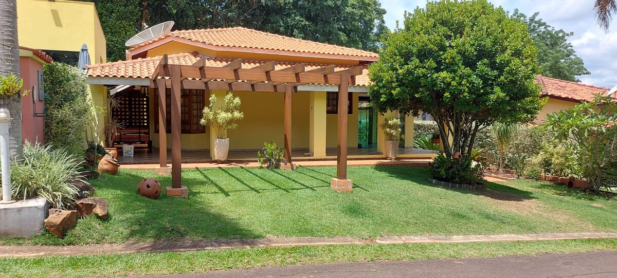 The exterior view features a charming house with a terracotta roof and a wooden pergola in the foreground. Lush green grass surrounds the property, with well-maintained shrubs and potted plants adding to the inviting atmosphere.