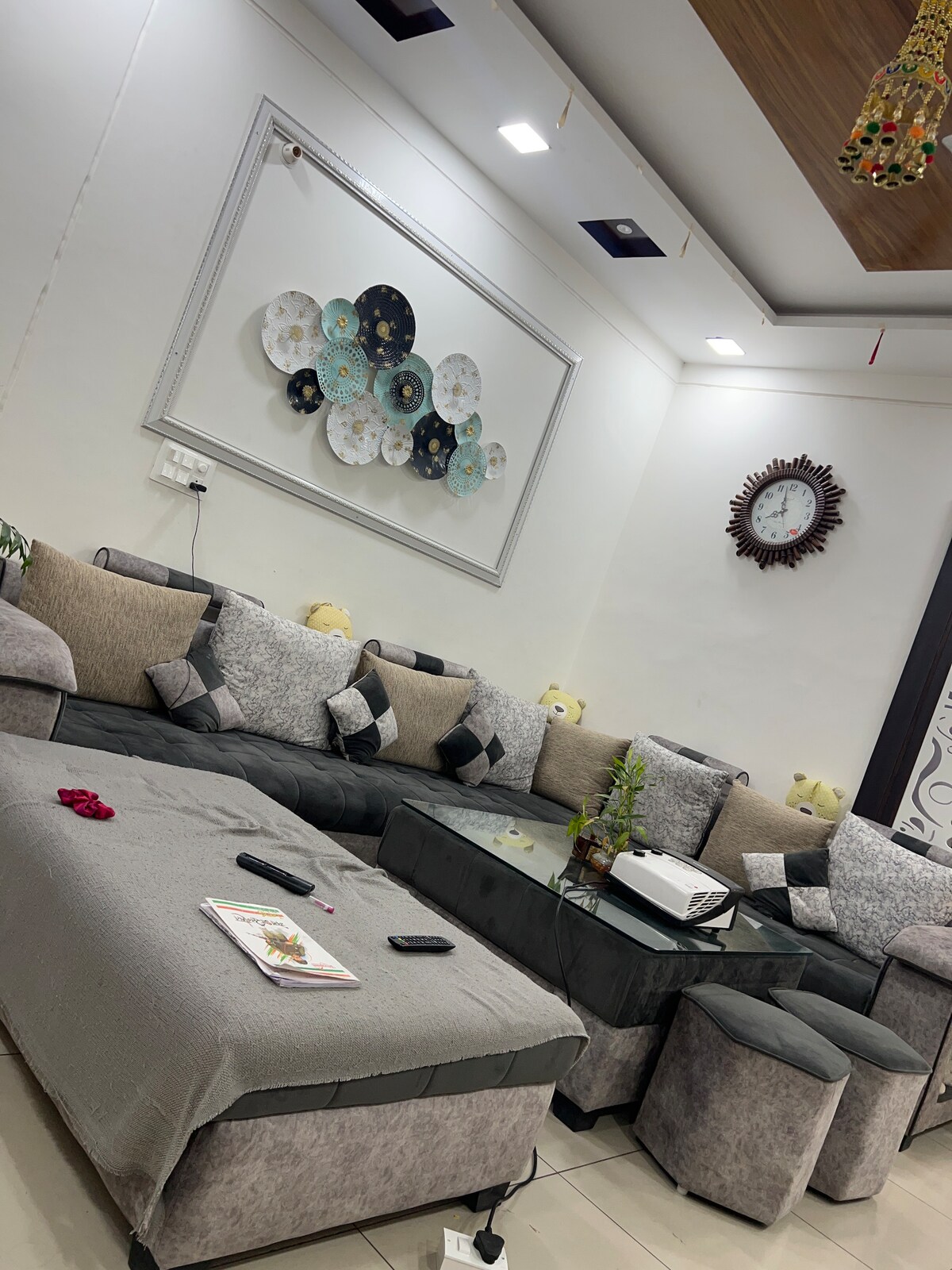 A comfortable seating area features a spacious sectional sofa with an array of decorative pillows. A low glass table sits at the center, surrounded by small stools. Wall art depicting colorful plates enhances the space, while soft lighting provides a warm ambiance.