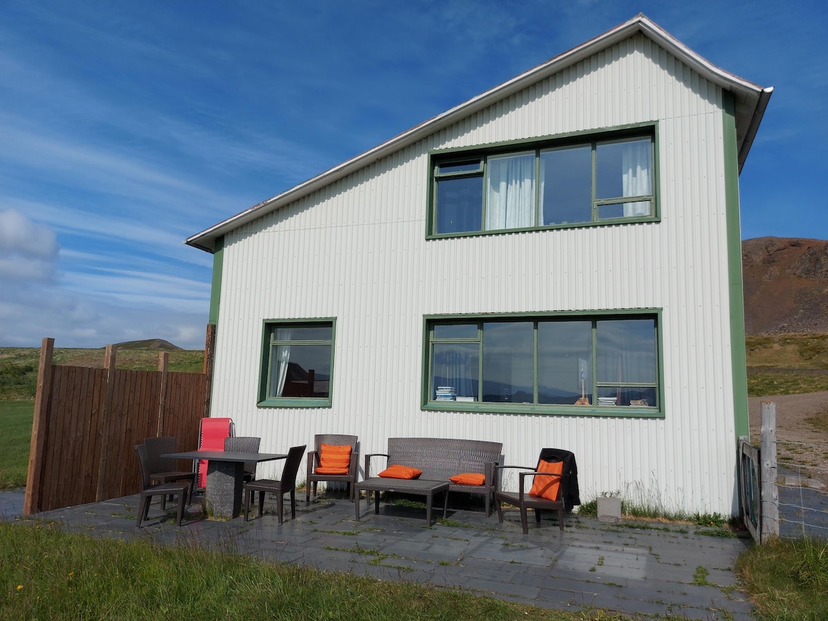 The exterior of the farmhouse presents a two-story structure with a light-colored facade. A covered patio area features outdoor seating, including a grey wicker couch and chairs with orange cushions, inviting relaxation while enjoying the surrounding landscape.