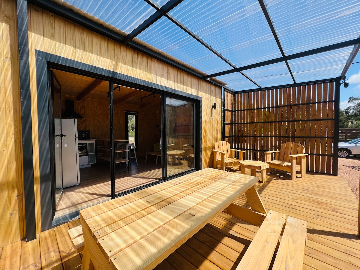An exterior deck features a wooden dining table, surrounded by lounge chairs. Large sliding glass doors provide a view of the interior seating area and kitchen. A covered roof allows for natural light while protecting the space from the elements.