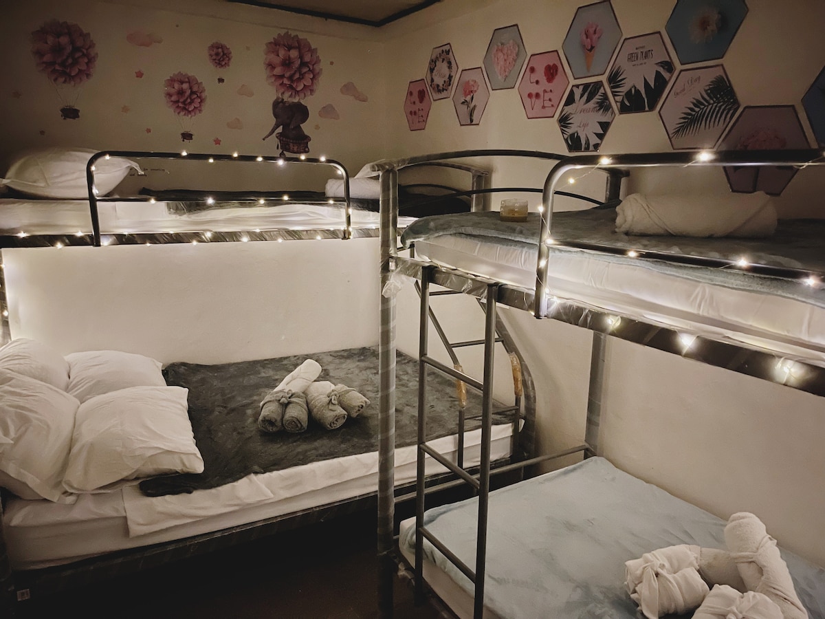 A shared sleeping area features two sets of bunk beds with soft bedding and multiple pillows. Decorative wall art, including floral patterns and hexagonal shapes, enhances the space. Warm string lights outline the bunk frames, providing a soft glow throughout the room.
