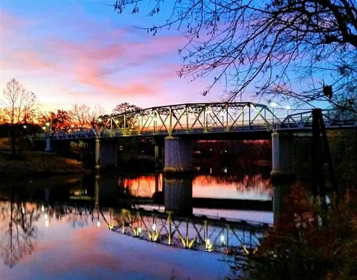 A metallic bridge spans across a calm river, reflecting vibrant colors of the sunset in the water. Surrounding trees and landscape are silhouetted against a backdrop of pastel pink and blue skies, creating a serene atmosphere.