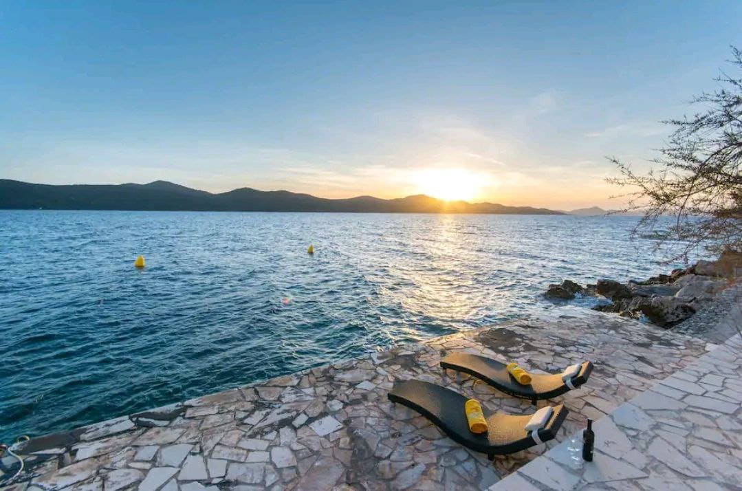 Two sun loungers are positioned on a stone pier at the water's edge, overlooking a vast expanse of shimmering sea. The sun is setting on the horizon, casting warm light across the water and illuminating the distant mountains.