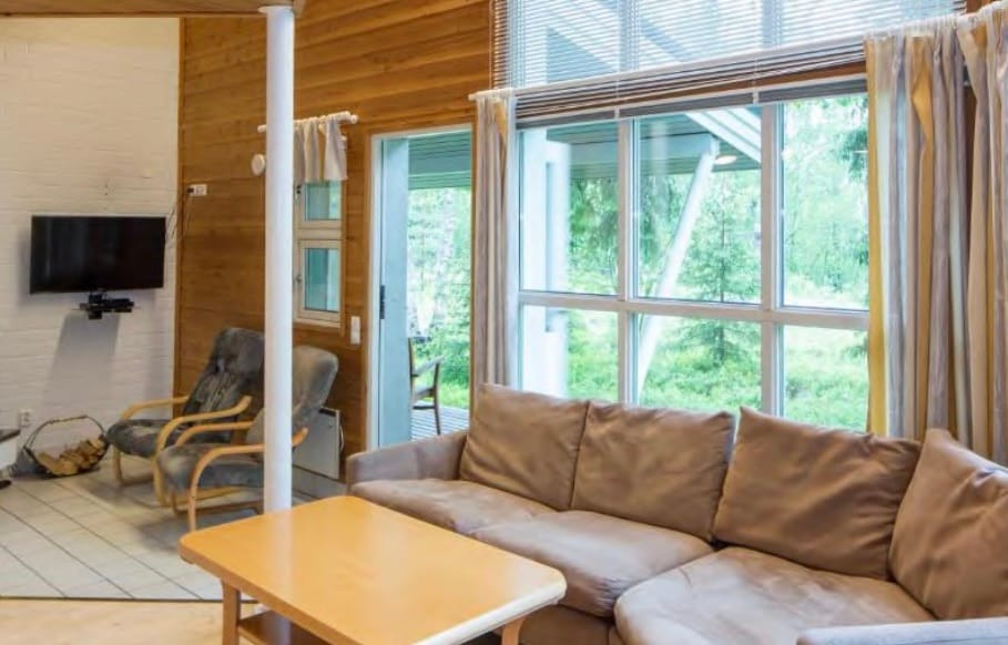 A bright and airy living area features large windows that offer views of the surrounding greenery. A comfortable sectional sofa and a wooden coffee table are positioned on a tiled floor, while two armchairs provide additional seating. Natural light floods the space, creating an inviting atmosphere.