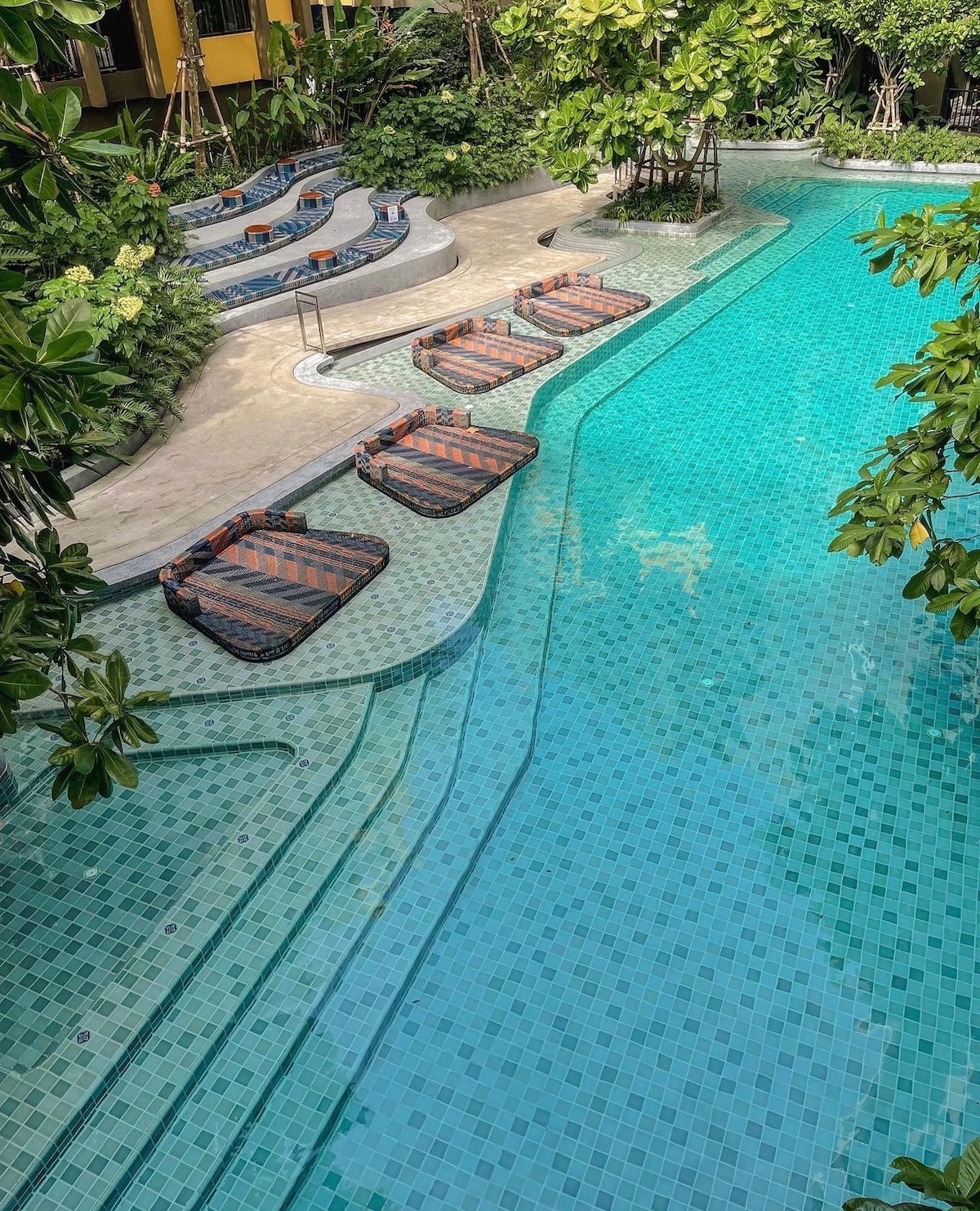 A spacious swimming pool is surrounded by lush greenery and tropical plants. Several lounging chairs with cushioned covers are positioned along the pool's edge. The water reflects shades of blue, while a small whirlpool area is visible in the background.