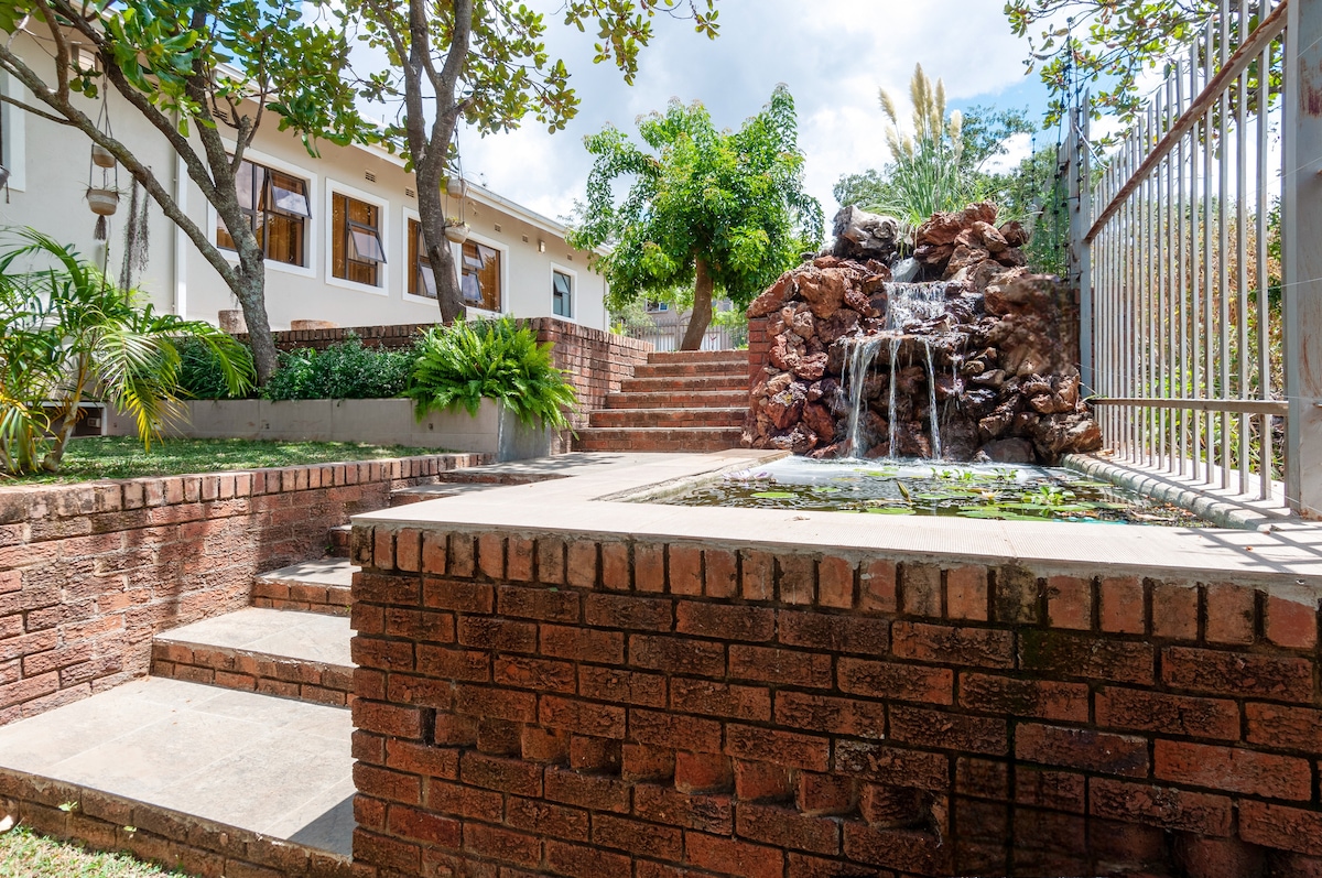 A tranquil outdoor area features a lush garden with natural stone steps leading to a serene waterfall and pond. Brick walls frame the space, providing structure and depth, while surrounding trees offer shade and greenery.
