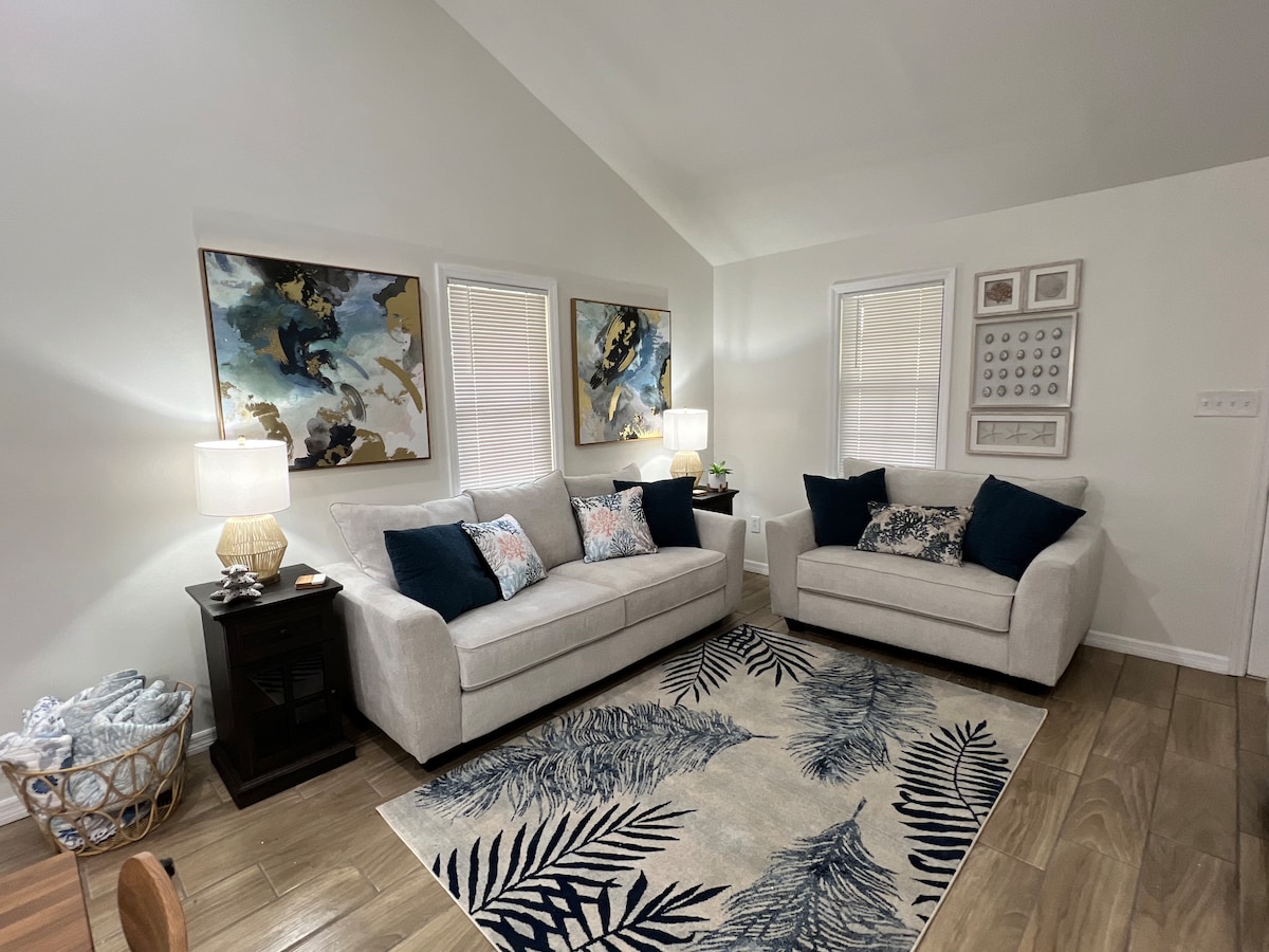 A well-lit living area features two light-colored sofas adorned with navy blue and patterned pillows. A decorative plant is placed on a side table, while abstract artwork hangs on the walls. A basket of blankets is positioned near the seating, enhancing comfort.