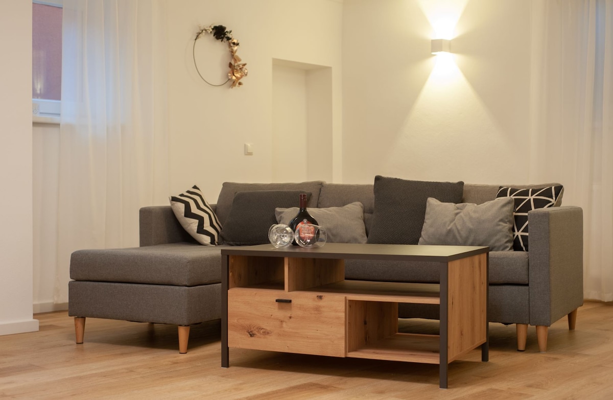 A modern living area features a sectional sofa in gray upholstery, adorned with decorative pillows in various patterns. A wooden coffee table with an open shelf enhances the space, while ambient lighting creates a soft atmosphere against the light walls.