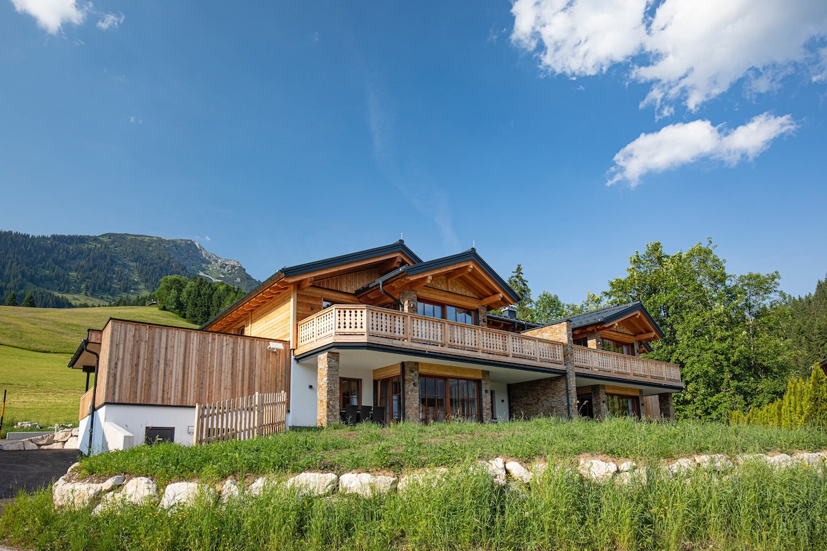 The exterior of the two-story chalet is showcased against a clear blue sky, featuring wooden balconies and natural stone elements. Lush greenery surrounds the property, while rolling hills and mountains provide a scenic backdrop.